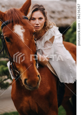 A woman in a white sundress riding a horse near a farm 96236267