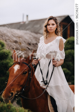 A woman in a white sundress riding a horse near a farm 96236277