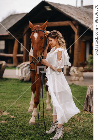 Beautiful girl in a white sundress next to a horse on an old ranch Beautiful girl in a white sundress next to a horse on an old ranch 96236370