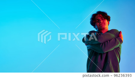 Portrait of young man with curly hair posing, hugging himself isolated over blue background in neon light. Self-love. Flyer 96236393