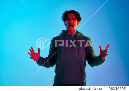 Portrait of young man with curly hair posing, emotively shouting isolated over blue background in neon light. Concept of emotions and lifestyle 96236395