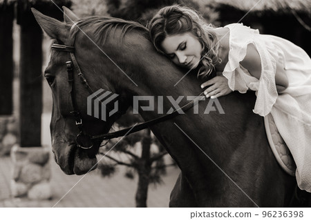 A woman in a white sundress riding a horse near a farm. black and white photo 96236398