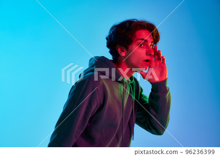 Portrait of young man with curly hair posing with hand to mouth, whispering secrets isolated over blue background in neon light. News 96236399