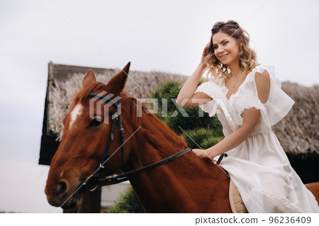 A woman in a white sundress riding a horse near a farm 96236409