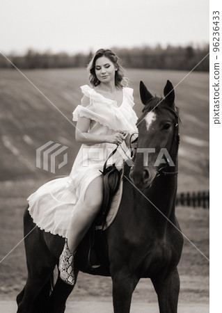 A woman in a white sundress riding a horse in a field. black and white photo 96236433
