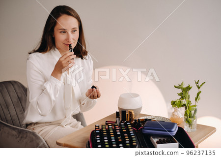The aromatherapist girl is sitting in her office and holding a bottle of aromatic oils. there are essential oils on the table The aromatherapist girl is sitting in her office and holding a bottle of aromatic oils. there are essential oils on the table 96237135