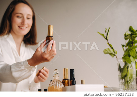 The aromatherapist girl is sitting in her office and holding a bottle of aromatic oils. there are essential oils on the table The aromatherapist girl is sitting in her office and holding a bottle of aromatic oils. there are essential oils on the table 96237138