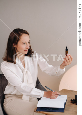 The aromatherapist girl is sitting in her office and holding a bottle of aromatic oil in her hands and writing something down. there are essential oils on the table The aromatherapist girl is sitting in her office and holding a bottle of aromatic oil in her hands and writing something down. there are essential oils on the table 96237174