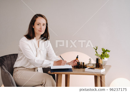 The aromatherapist girl is sitting in her office and holding a bottle of aromatic oil in her hands and writing something down. there are essential oils on the table 96237175