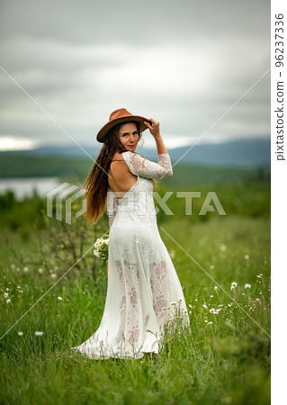 A middle-aged woman in a white dress and brown hat stands on a green field and holds a basket in her hands with a large bouquet of daisies. In the background there are mountains and a lake. A middle-aged woman in a white dress and brown hat stands on a green field and holds a basket in her hands with a large bouquet of daisies. In the background there are mountains and a lake. 96237336