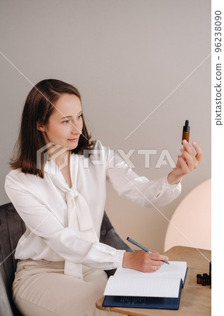 The aromatherapist girl is sitting in her office and holding a bottle of aromatic oil in her hands and writing something down. there are essential oils on the table The aromatherapist girl is sitting in her office and holding a bottle of aromatic oil in her hands and writing something down. there are essential oils on the table 96238090