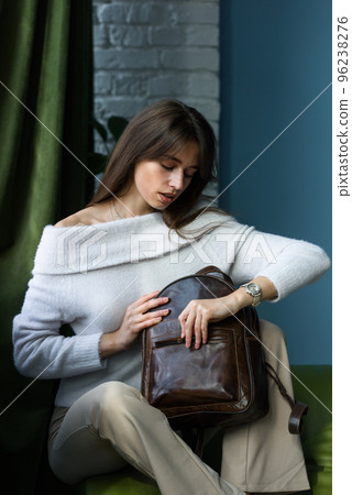 beautiful slender girl in beige pants and olive sweater posing while sitting on a green sofa with a small shiny brown leather backpack 96238276