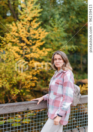 Portrait Of Cute Young Woman In Casual Wear In Autumn, Standing On Bridge Against Background Of An Autumn Park And River. Pretty Female Walking In Park In Golden Fall. Copy Space. Smiling Girl In The Portrait Of Cute Young Woman In Casual Wear In Autumn, Standing On Bridge Against Background Of An Autumn Park And River. Pretty Female Walking In Park In Golden Fall. Copy Space. Smiling Girl In The 96239415