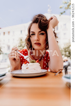 Charming woman in a restaurant, cafe on the street. She sits at the table and eats a cake with a fork. Dressed in a red sundress with white polka dots. Charming woman in a restaurant, cafe on the street. She sits at the table and eats a cake with a fork. Dressed in a red sundress with white polka dots. 96241215