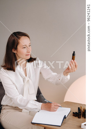The aromatherapist girl is sitting in her office and holding a bottle of aromatic oil in her hands and writing something down. there are essential oils on the table 96241241