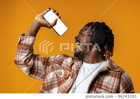 African guy in headphones singing song while listening music on smartphone on yellow background African guy in headphones singing song while listening music on smartphone on yellow background 96243263