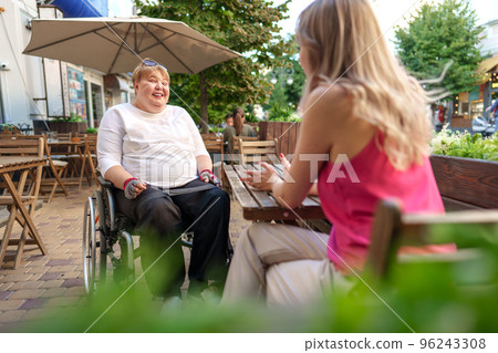 Mother with disability in wheelchair talking to her daughter while sitting at the table in cafe Mother with disability in wheelchair talking to her daughter while sitting at the table in cafe 96243308