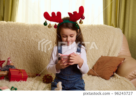 Beautiful Caucasian little child girl in blue casual denim overalls and funny deer antler hoop, holding a cute Christmas present in her hands, sitting on a comfortable sofa in the living room Beautiful Caucasian little child girl in blue casual denim overalls and funny deer antler hoop, holding a cute Christmas present in her hands, sitting on a comfortable sofa in the living room 96243727