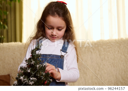 Close-up portrait of Caucasian pretty kid, beautiful little girl with long hair, enjoying decorating a Christmas tree with toys in the home interior. Christmastime. New Year preparations. Festive mood 96244157