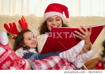 Adorable Caucasian little child girl, a lovely daughter listens to a fairy tale, sitting close to her loving mother in Santa hat, who is reading to her a book. Christmas mood. Winter holidays at home Adorable Caucasian little child girl, a lovely daughter listens to a fairy tale, sitting close to her loving mother in Santa hat, who is reading to her a book. Christmas mood. Winter holidays at home 96244831