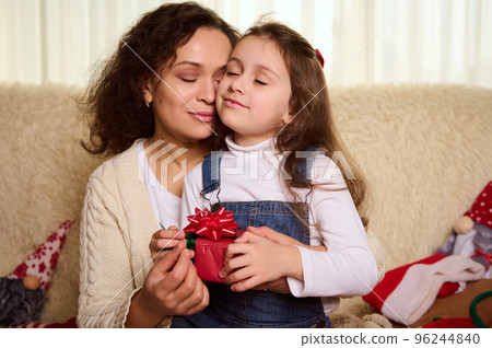 Moments of tenderness. Gorgeous dark-haired multi-ethnic pretty woman, a caring loving mom hugs her little daughter, while exchanging Christmas presents at home. Childhood. Happy family relationships 96244840