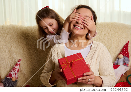 Pretty loving mother smiling a cheerful toothy smile, while her lovely daughter covers her eyes with her hands, offers her a Christmas present in stylish red box, sitting together on sofa at home 96244862