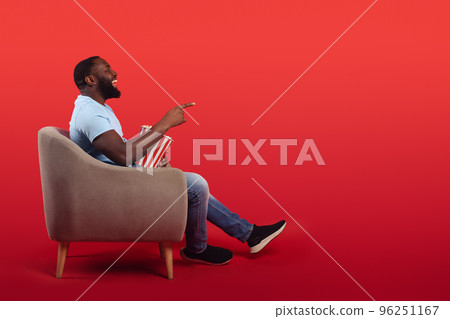 Amused televiewer sitting in the armchair at home while eating popcorns and watching a movie on red background Amused televiewer sitting in the armchair at home while eating popcorns and watching a movie on red background 96251167