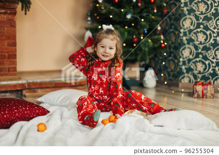 A cute little girl is sitting at the Christmas tree on Christmas morning with tangerines. Christmas time, gifts, festive mood. The concept of Christmas and New Year. Rear view A cute little girl is sitting at the Christmas tree on Christmas morning with tangerines. Christmas time, gifts, festive mood. The concept of Christmas and New Year. Rear view 96255304