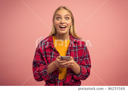 Blonde cute girl types a message with her smartphone. Happy and surprised expression face. Pink background Blonde cute girl types a message with her smartphone. Happy and surprised expression face. Pink background 96255568