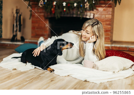 A smiling young woman is playing with a dog, sitting on the floor near the fireplace, on Christmas Eve. Christmas mood, Christmas holidays. The concept of Christmas and New Year A smiling young woman is playing with a dog, sitting on the floor near the fireplace, on Christmas Eve. Christmas mood, Christmas holidays. The concept of Christmas and New Year 96255865
