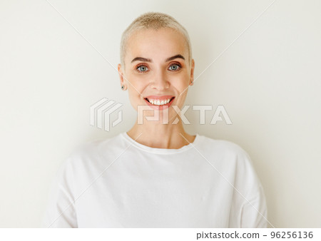 Beautiful woman with very short hair smiling at camera on blank wall Beautiful woman with very short hair smiling at camera on blank wall 96256136