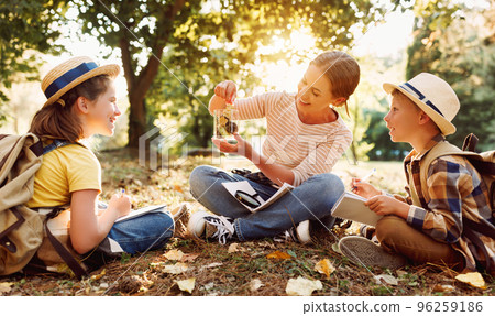 Happy mother and two children in with backpacks examining environment through magnifying glass in forest Happy mother and two children in with backpacks examining environment through magnifying glass in forest 96259186