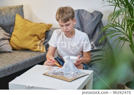 Schoolboy makes creative project in form of papier mache mountain. 96262478