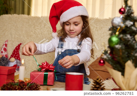 Beautiful little child girl in Santa's red hat, unpacking a cute gift box with surprise at Christmas. Winter holidays. Merry Christmas. Happy New Year. Festive mood at cozy home interior. December 25 96262781