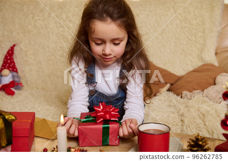 Charming Caucasian little girl with Christmas gift in the cozy home interior. A red cup of hot cocoa and lighted candle on journal table. Happy winter holidays. Childhood.New Year celebration concept 96262785