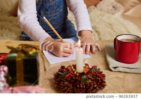 Selective focus on hand of unrecognizable child in denim overalls, drawing picture, writing letter to Santa, expecting the upcoming winter holidays. Christmas mood. Lighted candle on blur foreground 96263937