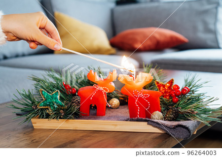 Female hand lighting a candle with a match in Christmas cozy composition for hygge home mood. Deer shape red candles, fresh green pine branches and Xmas decor on the coffee table. Selective focus. Female hand lighting a candle with a match in Christmas cozy composition for hygge home mood. Deer shape red candles, fresh green pine branches and Xmas decor on the coffee table. Selective focus. 96264003