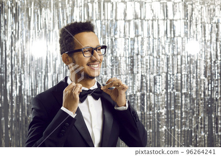 Happy handsome young ethnic man in tuxedo and stylish glasses adjusting his bow tie 96264241