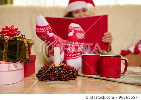 Selective focus on red socks with Christmas pattern, on blurred background of a pretty woman in Santa's hat reading book, relaxing on a comfortable sofa during New Years winter holidays. Copy ad space 96265657
