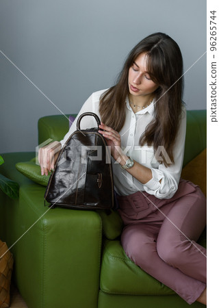 beautiful slender girl in burgundy pants and a white blouse posing while sitting on a green sofa with a small shiny brown leather backpack 96265744
