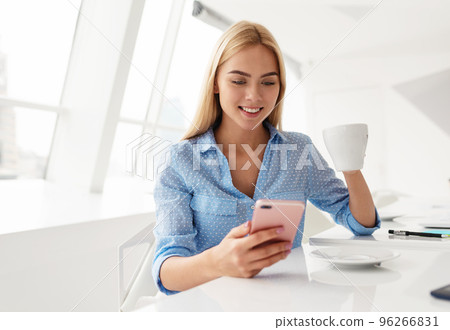 Smiling young woman with cellphone in hand while drinking coffee in a white room Smiling young woman with cellphone in hand while drinking coffee in a white room 96266831