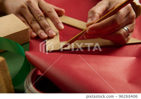 Close-up woman's hands using a wooden triangular ruler to draw on red gift paper, calculating the amount of packaging materials needed to wrap gifts for Christmas, New Year or any other holiday event Close-up woman's hands using a wooden triangular ruler to draw on red gift paper, calculating the amount of packaging materials needed to wrap gifts for Christmas, New Year or any other holiday event 96268406