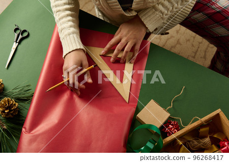 Top view woman's hands using a wooden triangular ruler to draw on red gift paper, calculating the amount of packaging materials needed to wrap gifts for Christmas, New Year or any other holiday event Top view woman's hands using a wooden triangular ruler to draw on red gift paper, calculating the amount of packaging materials needed to wrap gifts for Christmas, New Year or any other holiday event 96268417