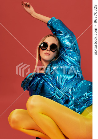 Young cute nice blonde girl in blue jacket sits in a fetal position in studio before camera, red background. Side view Young cute nice blonde girl in blue jacket sits in a fetal position in studio before camera, red background. Side view 96270328