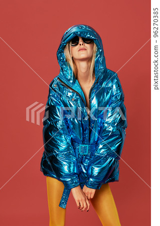 Blonde slim girl in blue down jacket covers her face from wind with hood 96270385