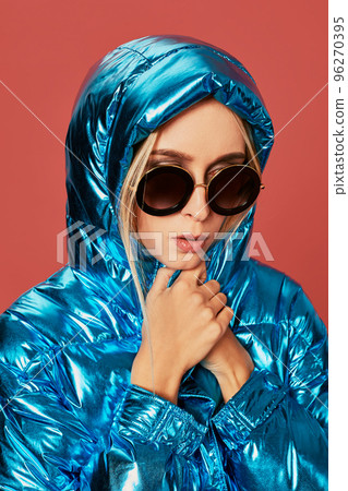 Blonde slim girl in blue down jacket covers her face from wind with hood 96270395