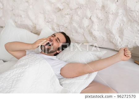 Cheerful millennial european man with stubble wakes up, yawns on white bed, enjoy cozy, comfort and lazy 96272570