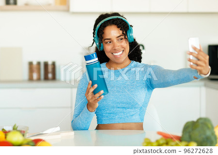 Happy african american sporty woman with bottle of water taking selfie on break from home workout, sitting in kitchen 96272628