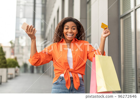 Glad excited young black woman with smartphone, packages and credit card raises hands up rejoices at great purchase Glad excited young black woman with smartphone, packages and credit card raises hands up rejoices at great purchase 96272646