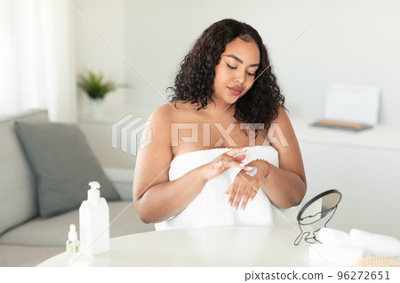 Black oversize woman applying moisturizing cream or lotion on her hands, sitting wrapped in towel at home, free space Black oversize woman applying moisturizing cream or lotion on her hands, sitting wrapped in towel at home, free space 96272651